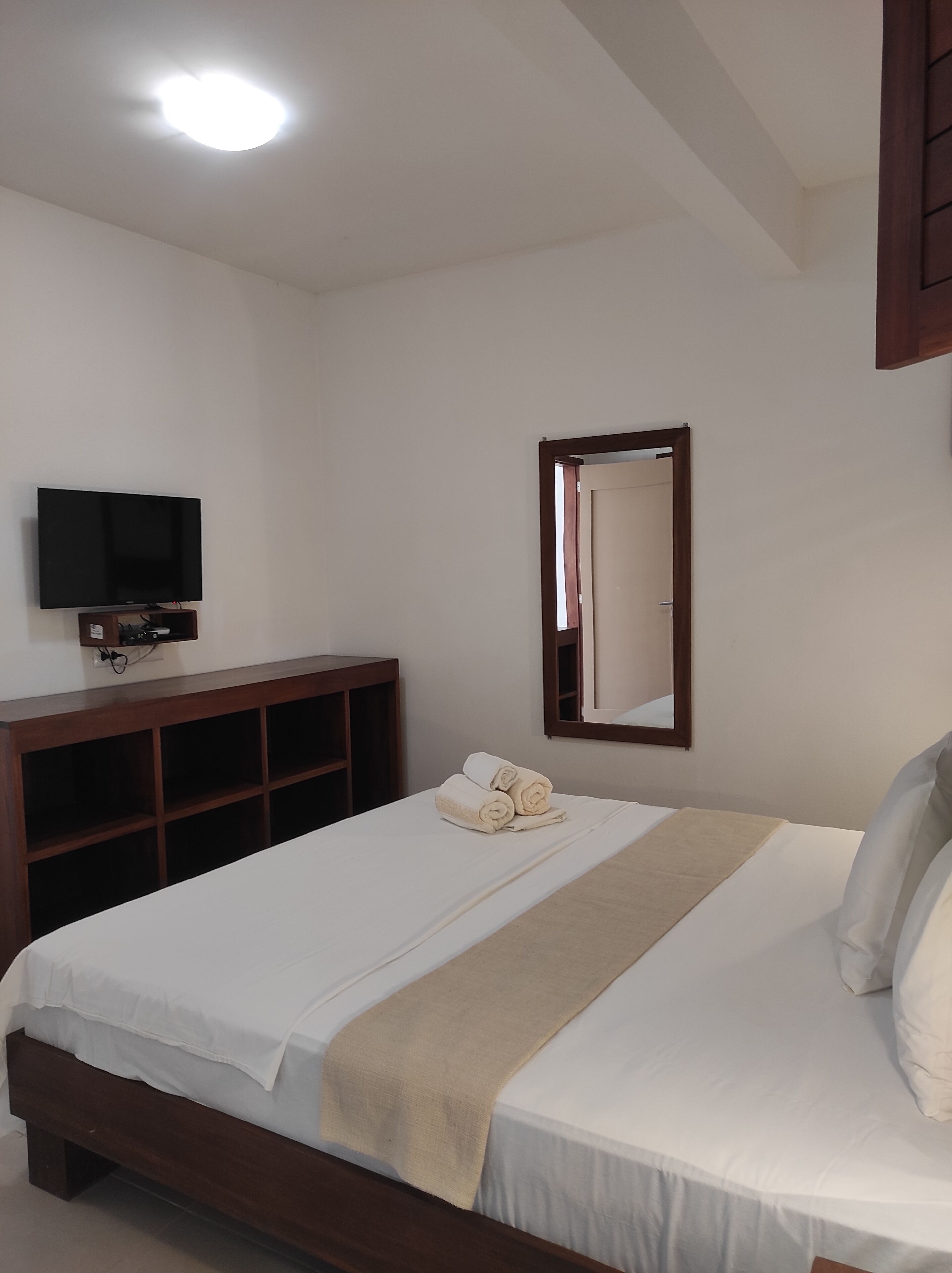 A bedroom with a bed, a TV mounted on the wall, a mirror, and a wooden shelf.