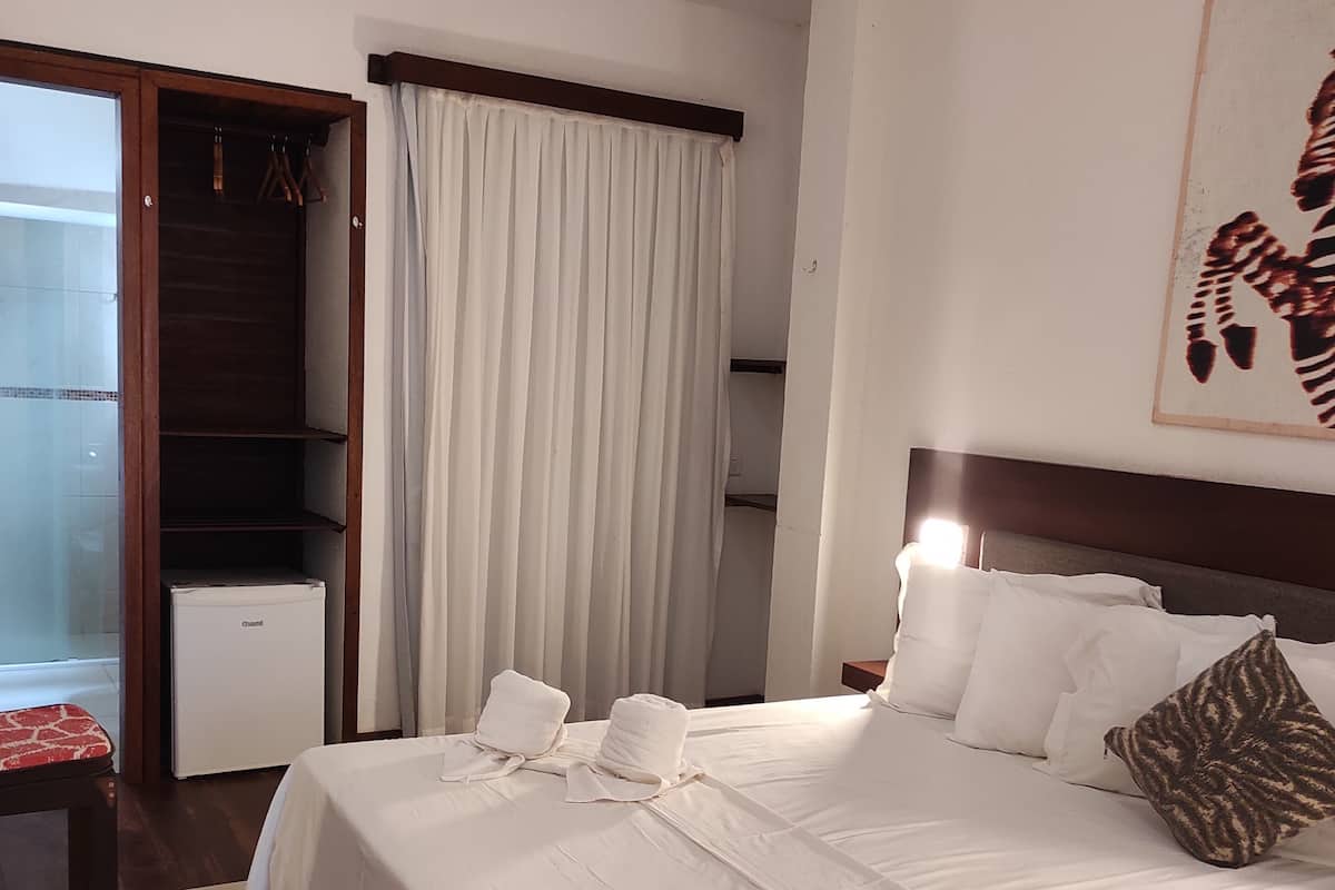 Una habitación de hotel con una cama grande, un armario, un refrigerador pequeño y una ventana con cortinas.