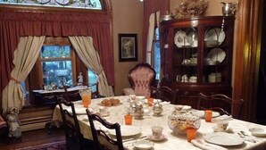 Une salle à manger avec une table ronde dressée pour quatre personnes, entourée de chaises en bois. Un buffet en porcelaine avec des portes en verre se trouve à droite, et une fenêtre avec des rideaux est visible au fond.