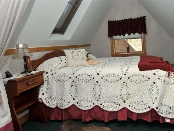 Ein Schlafzimmer mit schrägen Wänden, einem Bett mit Blumenmuster-Quilt, einem Nachttisch, einer Lampe und einem Fenster mit Verdunkelungsvorhang.