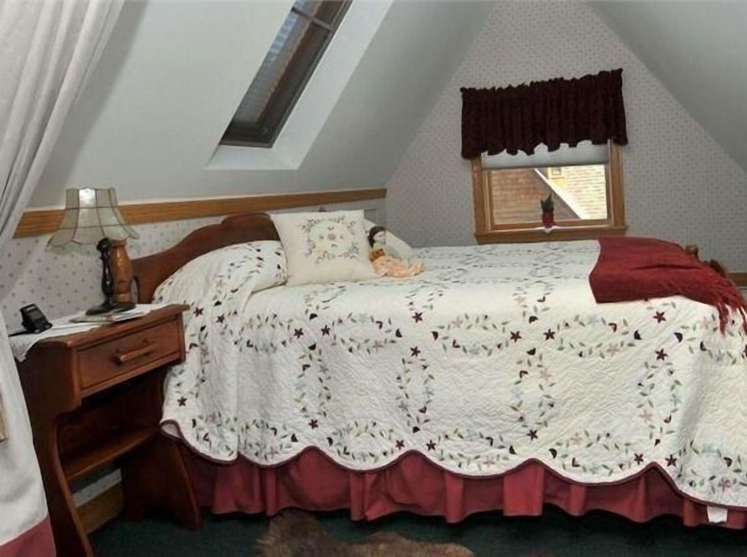 Un dormitorio con techo inclinado, estructura de cama de madera, mesita de noche, lámpara, ventana con cortina oscura y colcha con estampado floral.