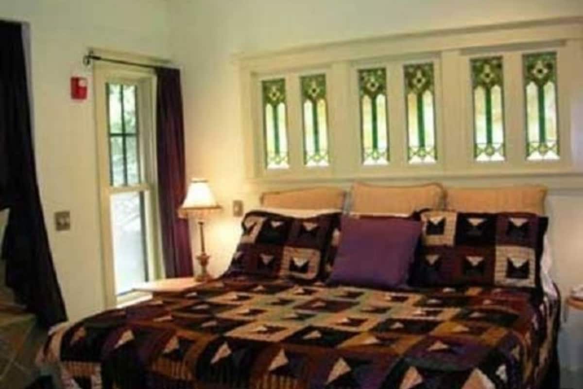 Quarto com cama, mesa de cabeceira, luminária e janela com detalhes decorativos.