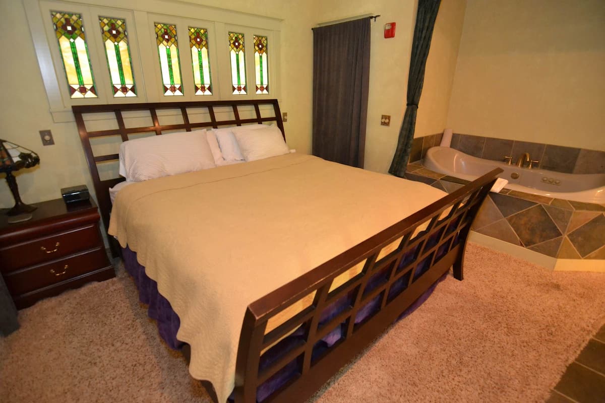 Quarto com cama, mesa de cabeceira, cômoda, janela com vitrais, banheira e chuveiro.