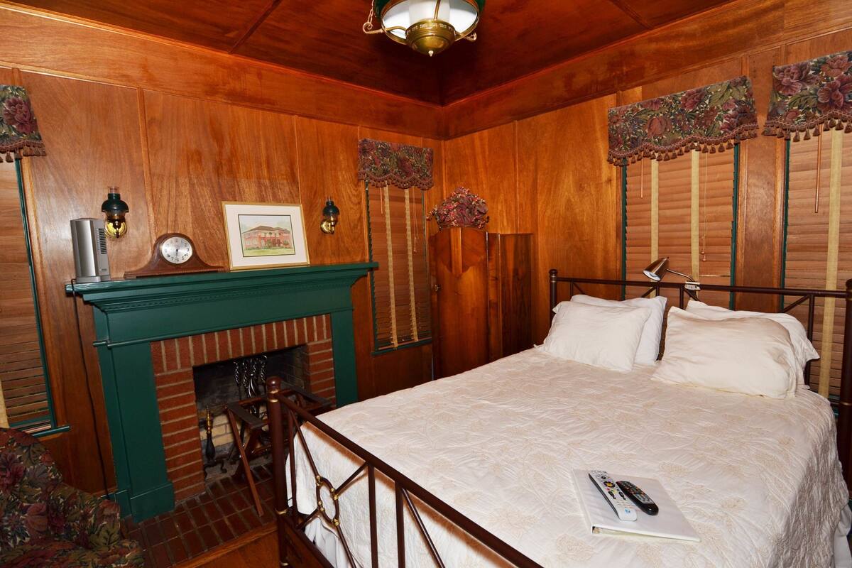 Quarto com teto de madeira, cama com roupa de cama branca, lareira e relógio na lareira.