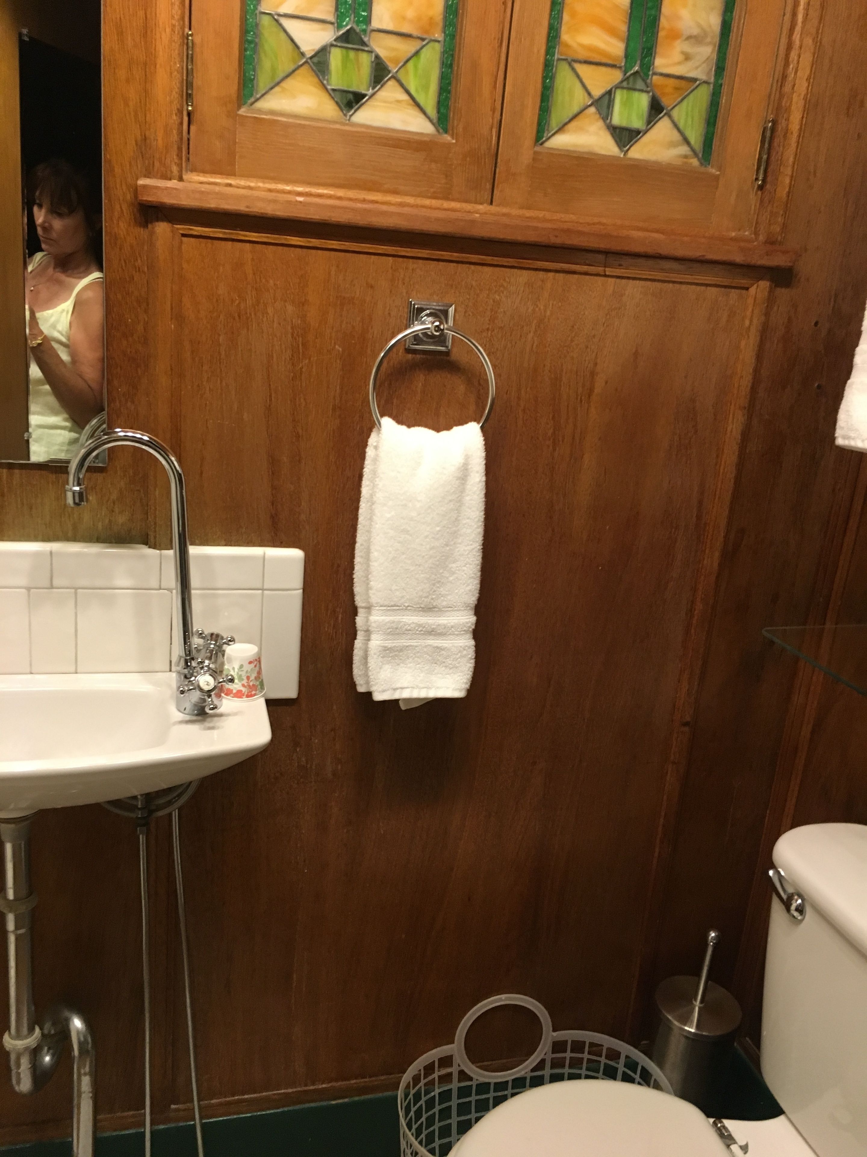 A bathroom with a wooden vanity, a white sink with a faucet, a glass cup, a white towel hanging on a metal ring, and a toilet.