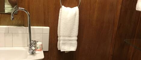 A bathroom with a wooden vanity, a white sink with a faucet, a glass cup, a white towel hanging on a metal ring, and a toilet.