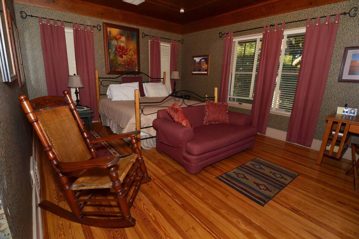 Quarto com poltrona de balanço de madeira, sofá roxo, cama com roupa de cama branca e piso de madeira.