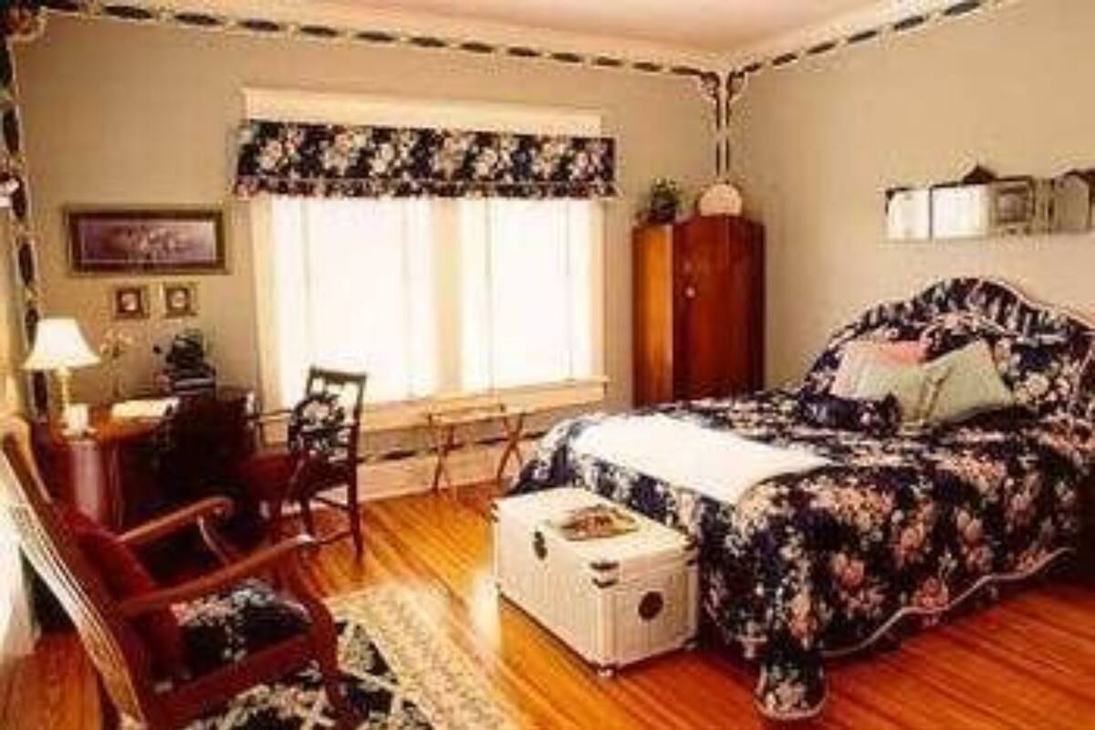Quarto com colcha florida, cómoda de madeira, secretária com candeeiro e janela com cortinas.