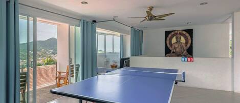 A spacious room with a ping pong table, ceiling fans, large windows, and a mural on the wall.