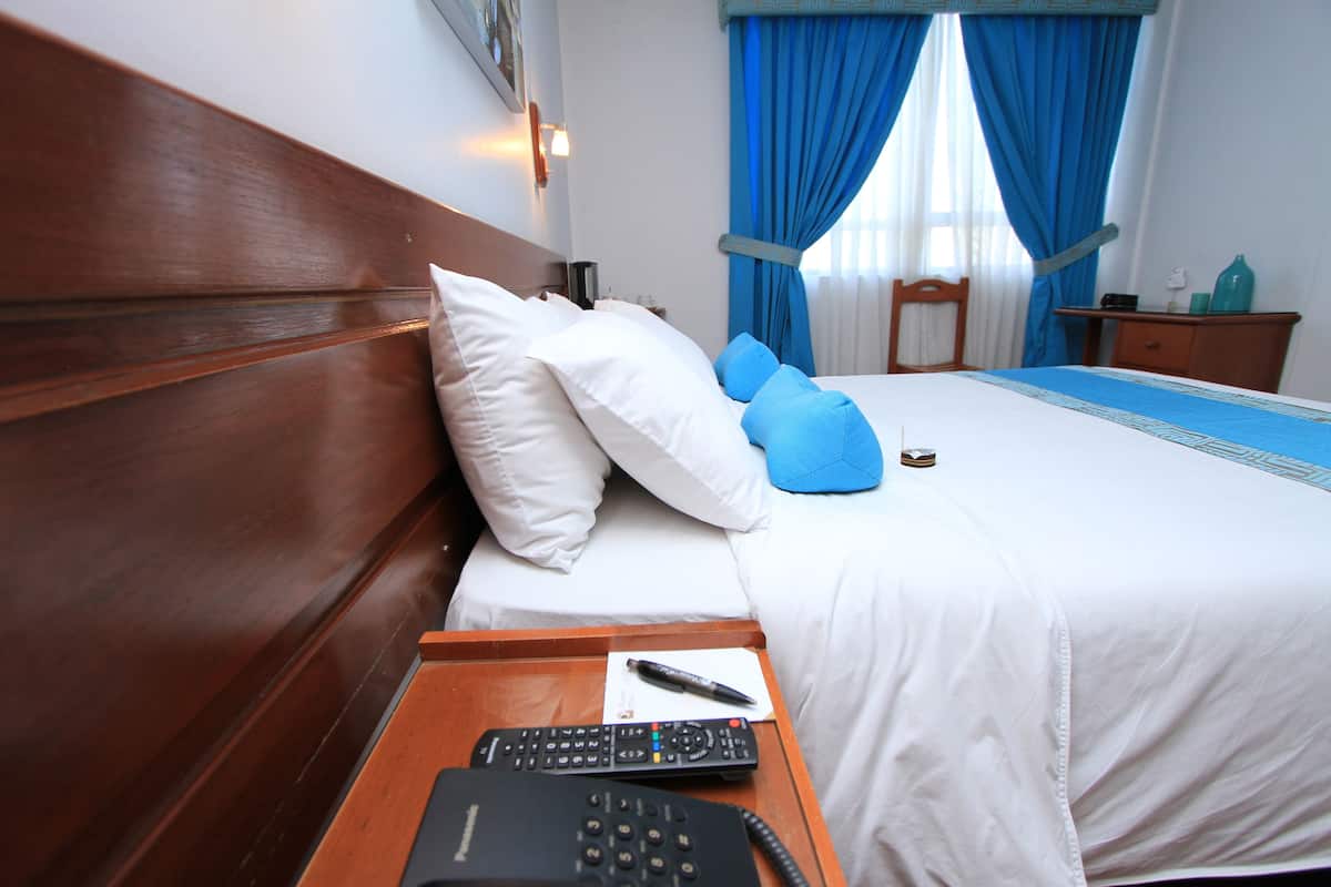 Habitación de hotel con cabecera de madera, cama con ropa de cama blanca y azul, mesita de noche con un control remoto y un teléfono, y un velador con una lámpara y una botella.