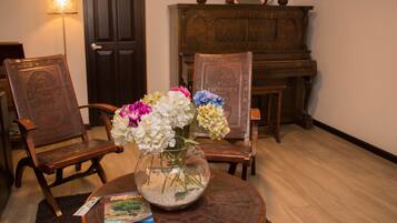 A room with a piano, two wooden chairs, a vase with flowers, and a lamp.