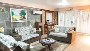 A living room with two sofas, a coffee table with magazines, a stone wall with a painting, and a door with curtains. - Hotel Pousada Ald'Mama (Gramado)