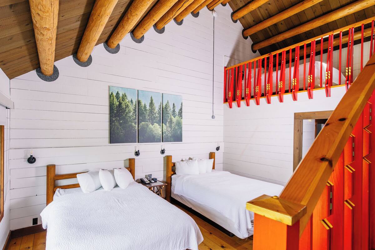 A room with two beds, wooden beams on the ceiling, a red railing, and a painting of a forest.