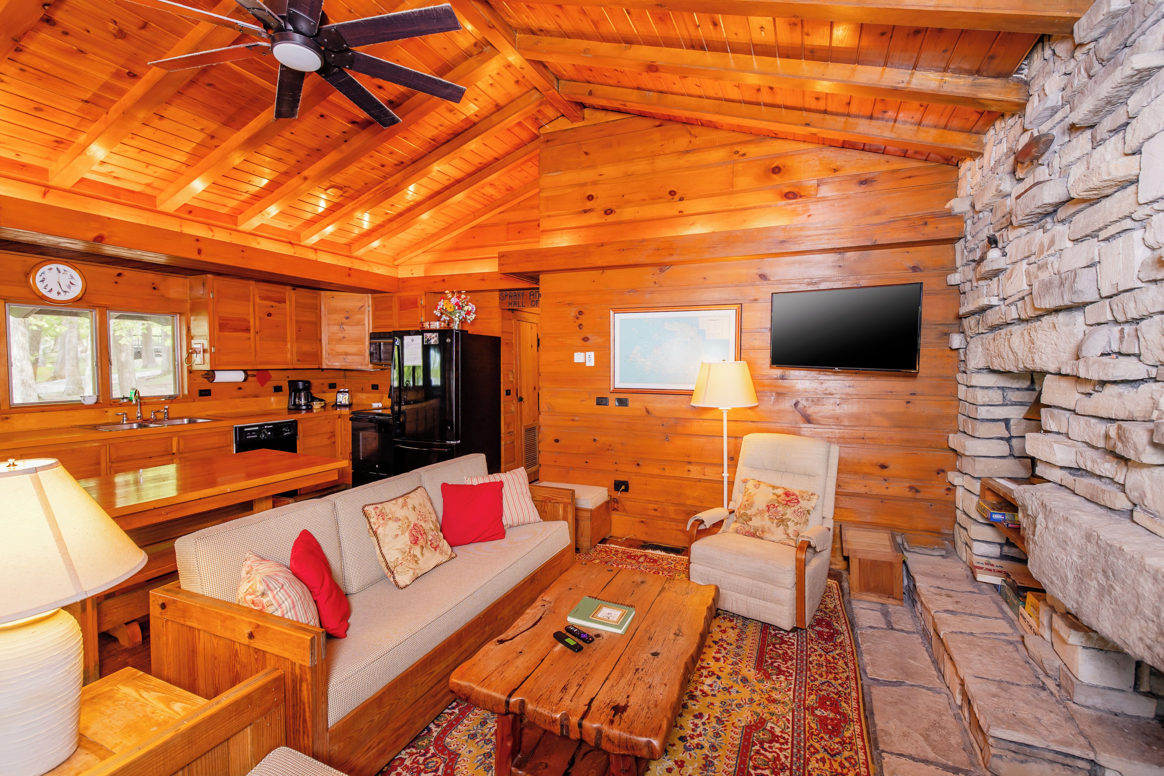 Un intérieur de chalet chaleureux avec des murs en bois, un mur d’accent en pierre, un canapé, un fauteuil, une table basse, un téléviseur fixé au mur et un ventilateur de plafond.