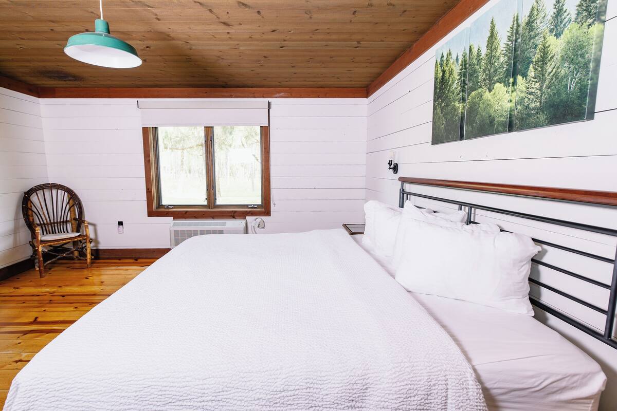 A bedroom with a large bed, wooden floor, a window, a chair, and a painting of trees.