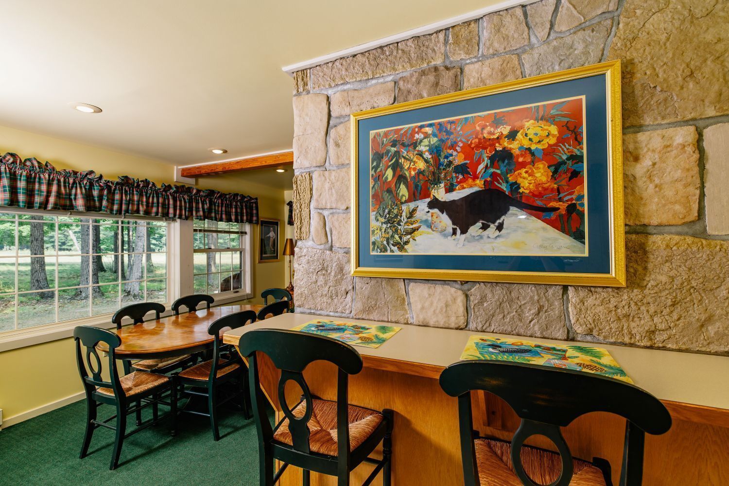 A dining area with a stone wall, a painting of a cat, and a view of trees through large windows.