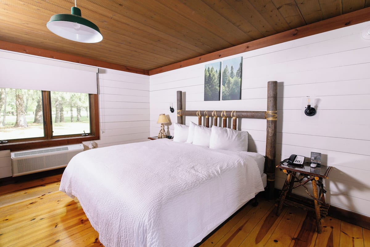 A bedroom with a large bed, wooden floor, a window with a view of trees, a wall-mounted heater, and a small bedside table with a lamp and a phone.