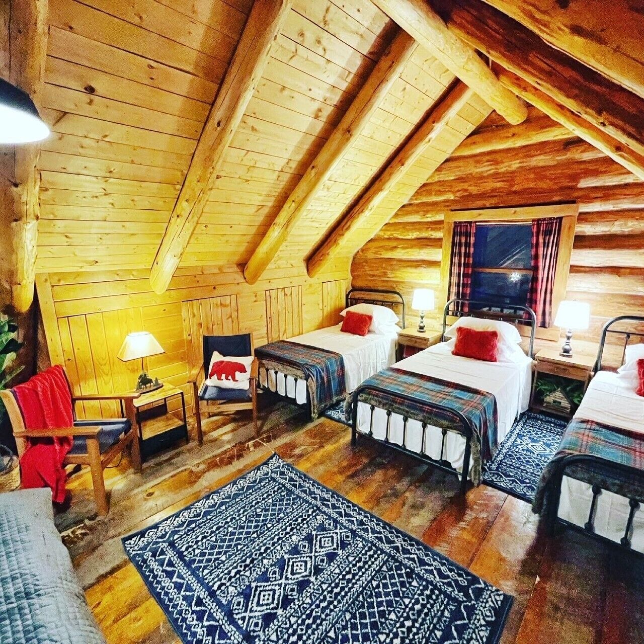 A cozy attic bedroom with wooden walls, two beds with plaid bedding, a small desk, and a chair.