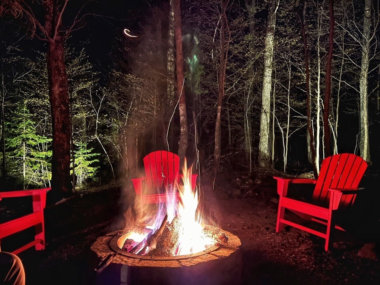Ein Lagerfeuer mit zwei roten Adirondack-Stühlen daneben, umgeben von einem Wald.