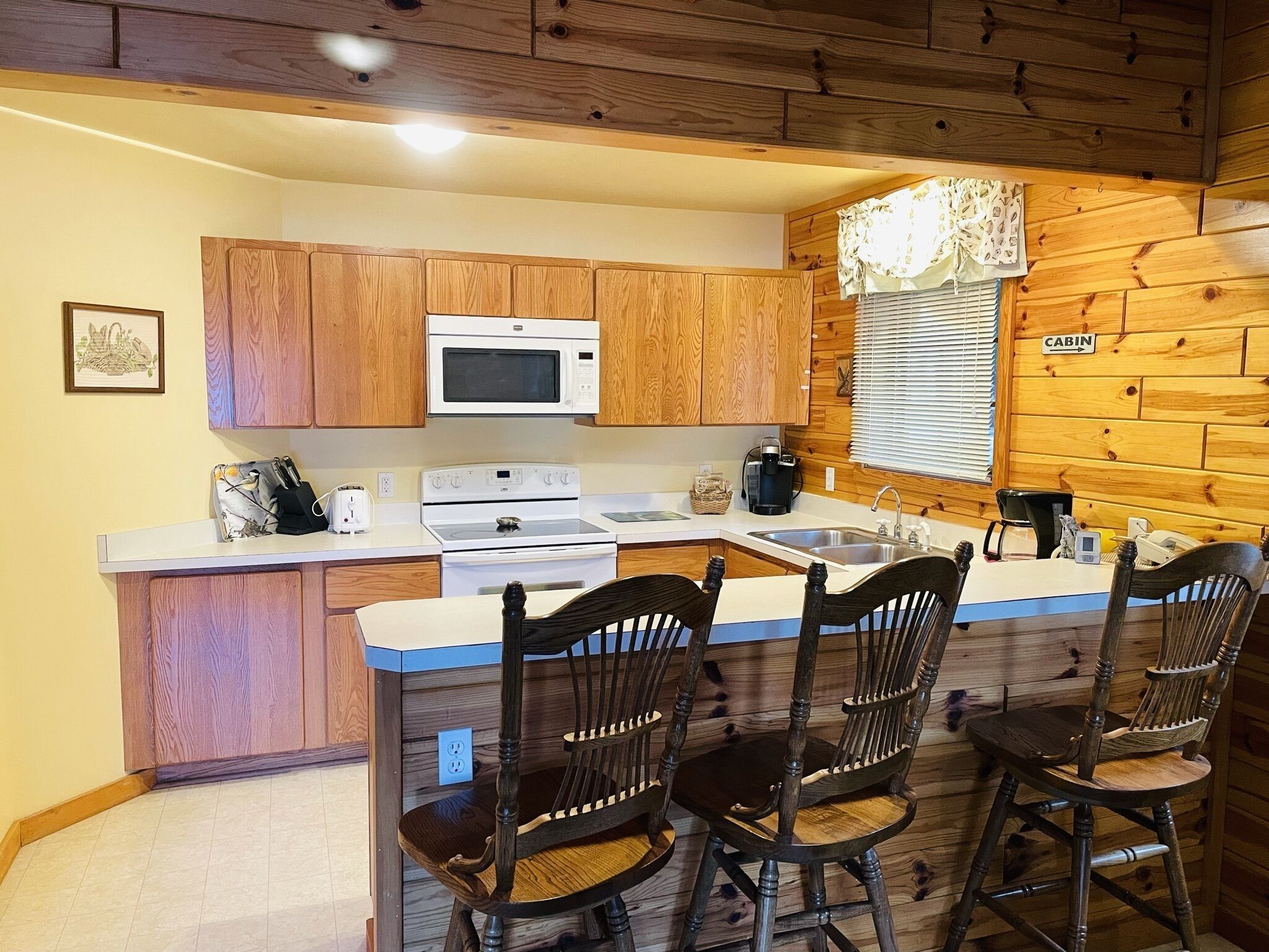 A compact kitchen with wooden cabinets, a microwave, stove, and a sink. There are four wooden chairs around a small table.