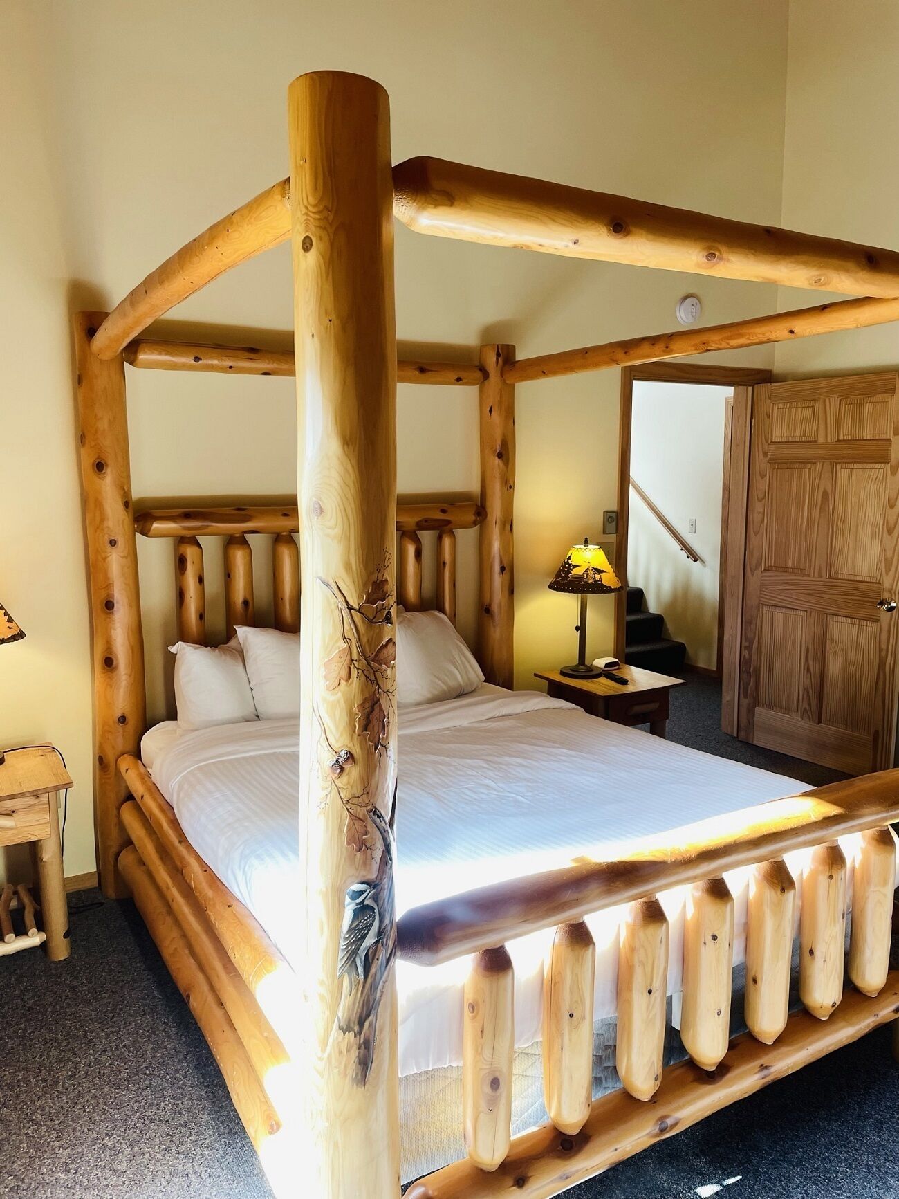 A wooden four-poster bed with a canopy, a nightstand with a lamp, and a wooden door in a room with carpeted flooring.