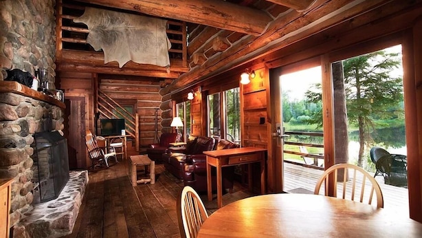 A rustic cabin interior with a stone fireplace, wooden furniture, and a large window overlooking a natural landscape. - Drummond Island Resort (Drummond Island)