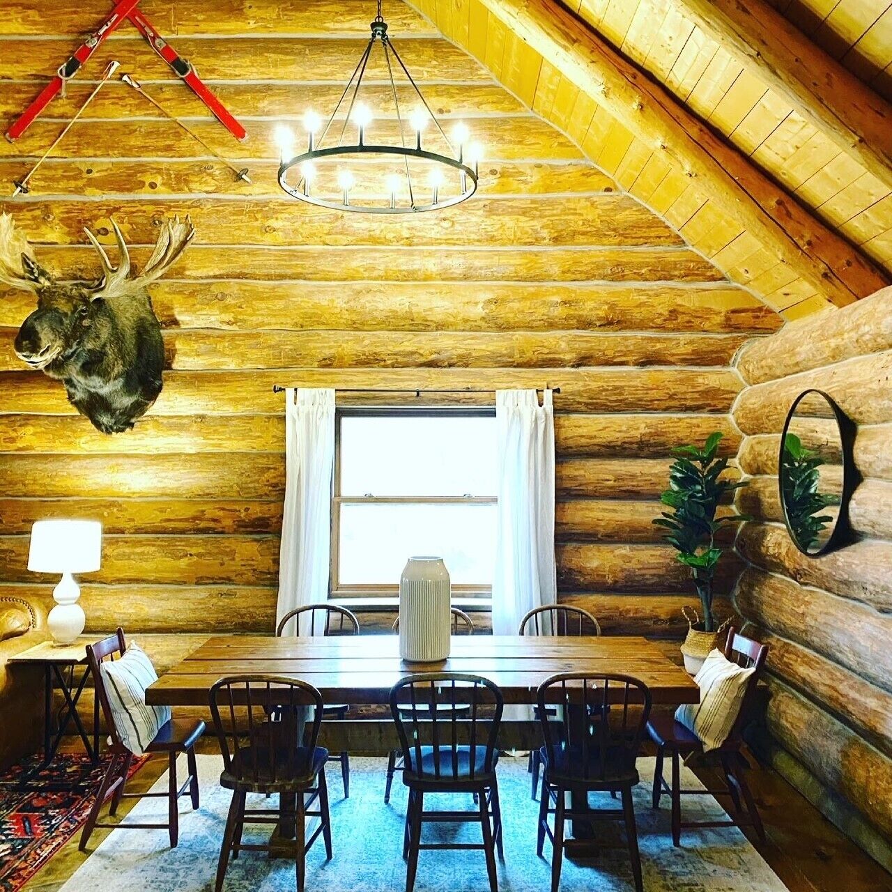 Interior de una cabaña de troncos con comedor, candelabro, cabeza de animal montada y ventana con cortinas.