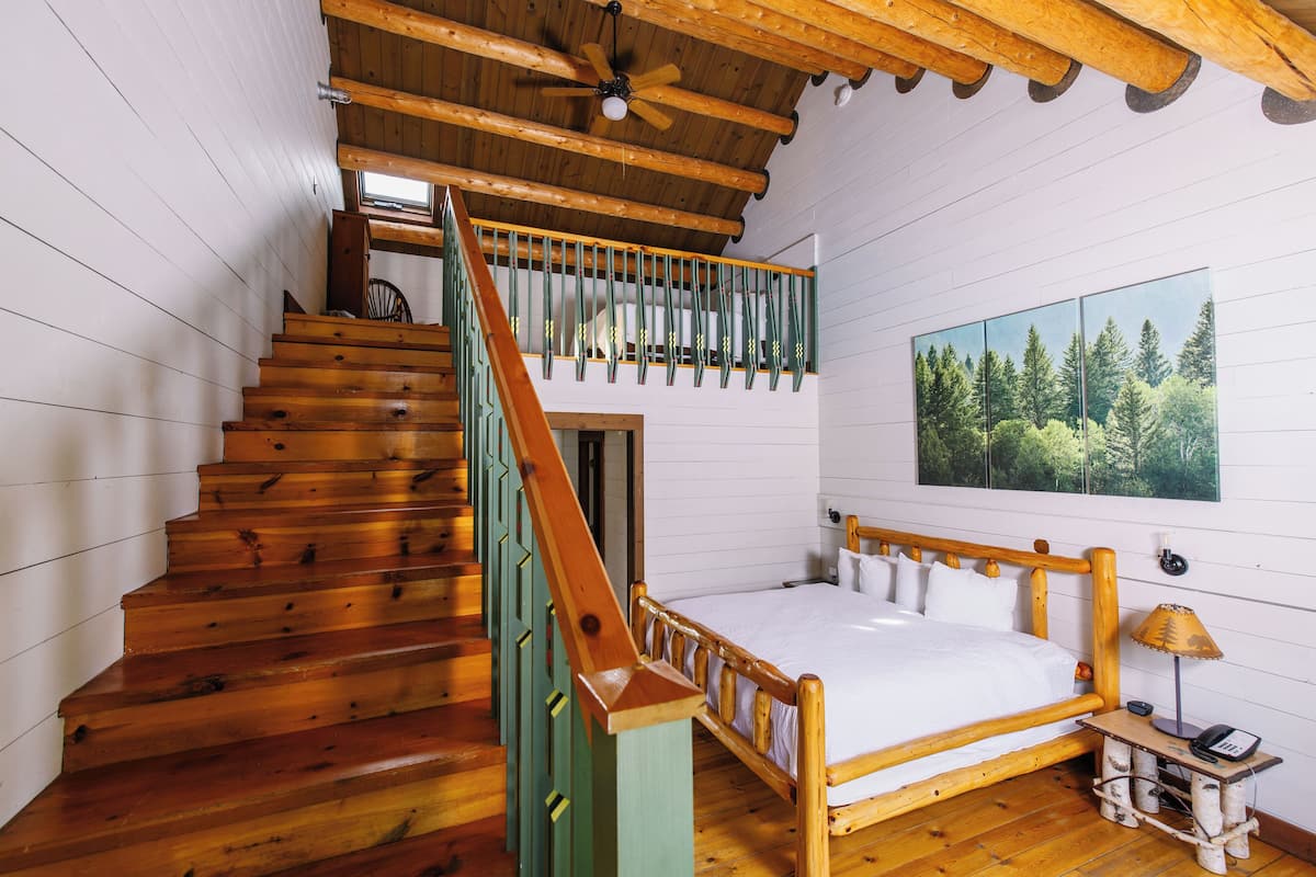 A wooden staircase leading to a loft bedroom with a bed and a painting of a forest.