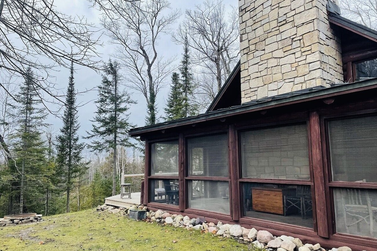 A cabin with large windows, a stone chimney, and a deck area.