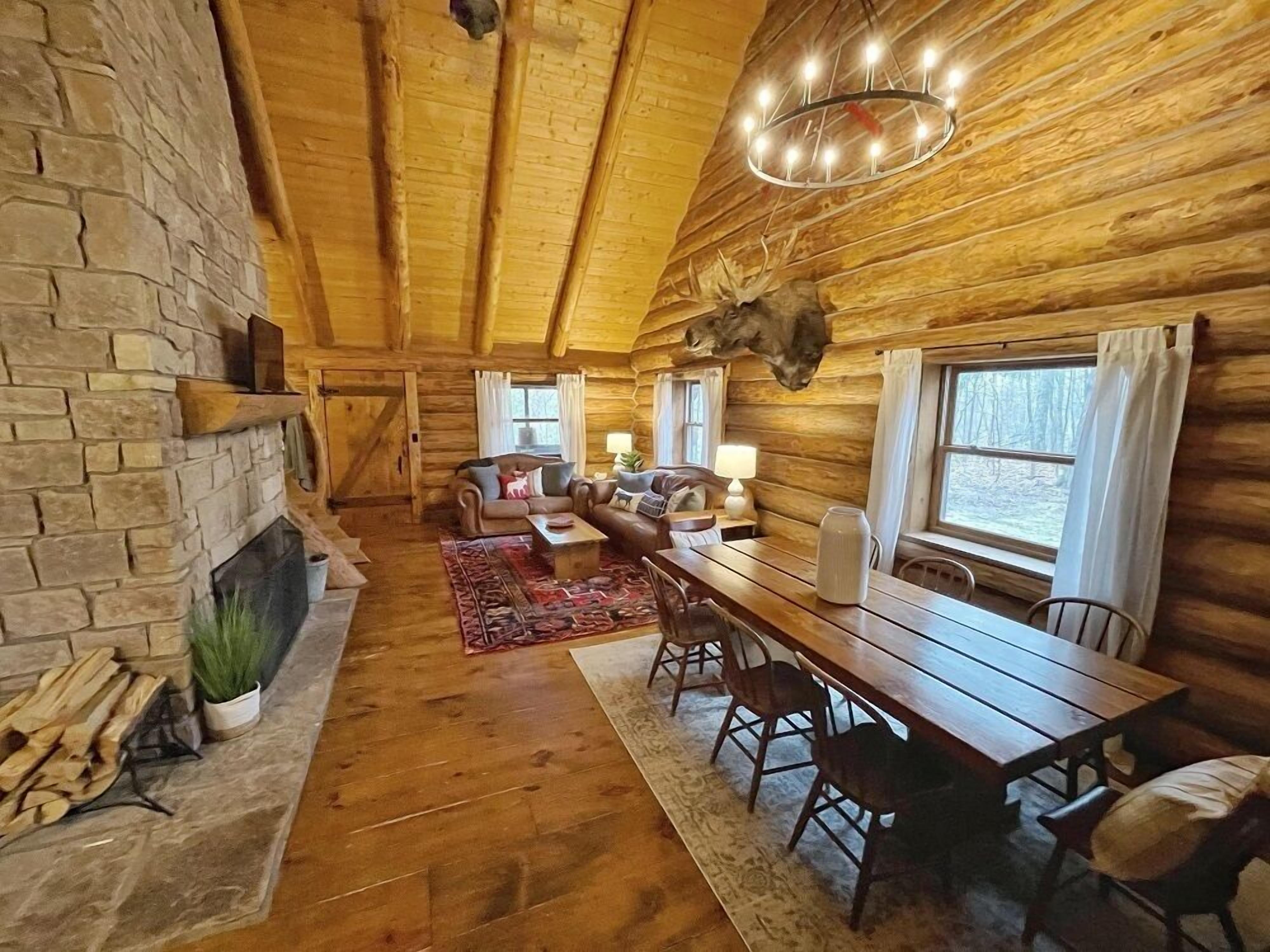 Interior de una cabaña de troncos con chimenea de piedra, una mesa de comedor de madera y una zona de estar acogedora.