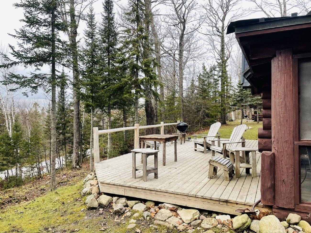 A wooden deck with Adirondack chairs and a grill, surrounded by trees and a view of a lake.