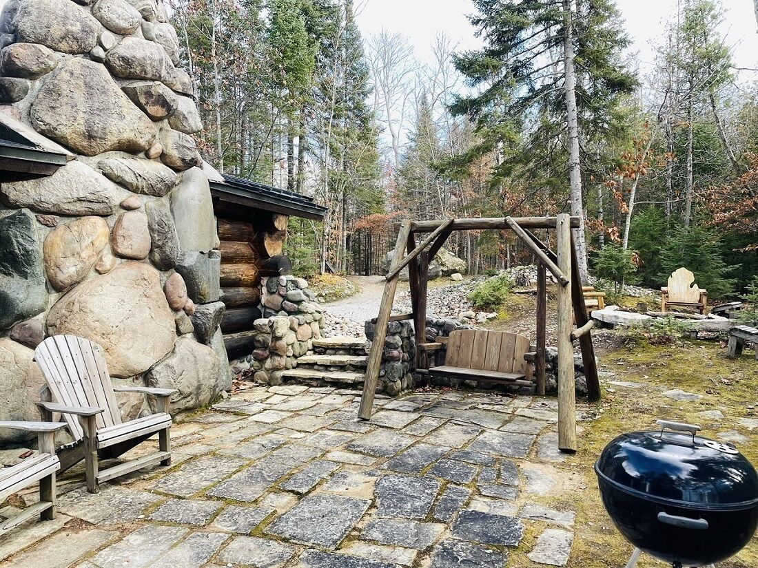 Metsäisellä alueella sijaitseva maalaishenkinen mökki, jossa on kiviseinä, puinen riippukeinu ja grilli.