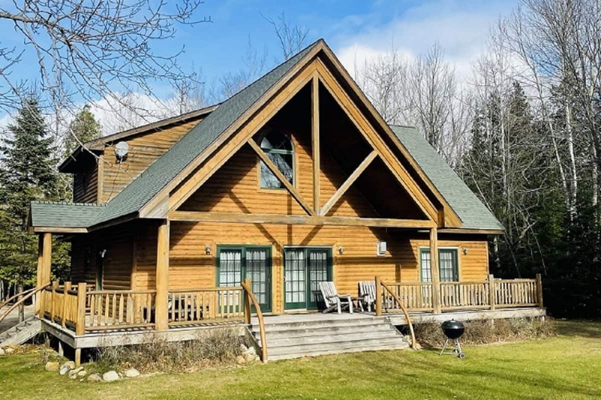 A log cabin with a prominent triangular roof, a porch with steps, and a grill in the yard.