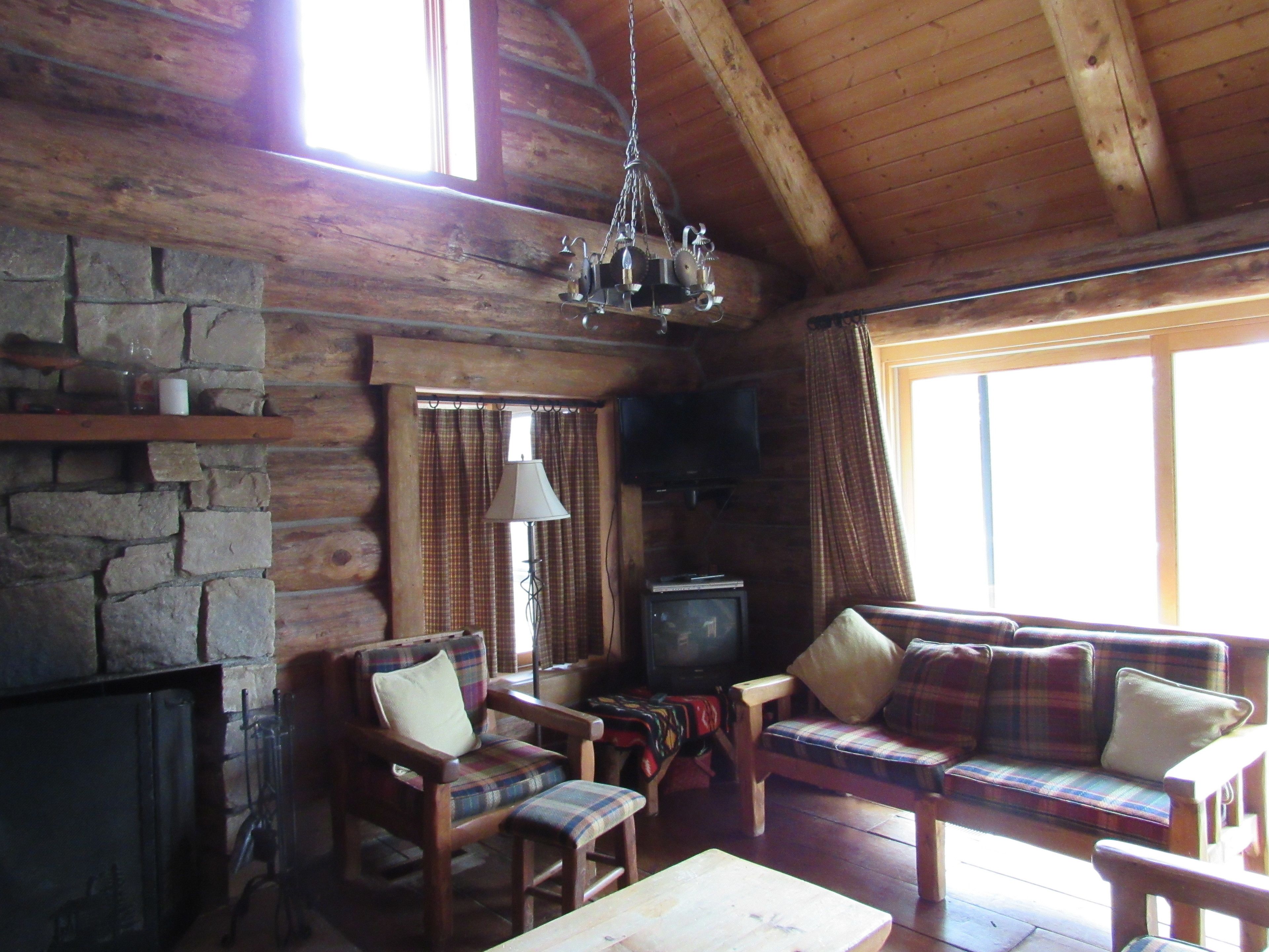 L’intérieur d’un chalet rustique, avec une cheminée en pierre, des meubles en bois, un téléviseur et une grande fenêtre agrémentée de rideaux.
