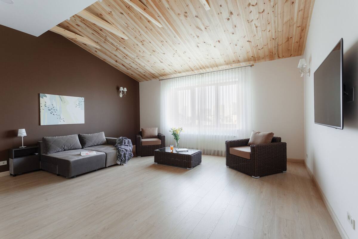 Una sala de estar moderna con techo de madera, un televisor de pantalla plana grande, dos sofás, una mesa de centro y un ventanal con cortinas de gasa.