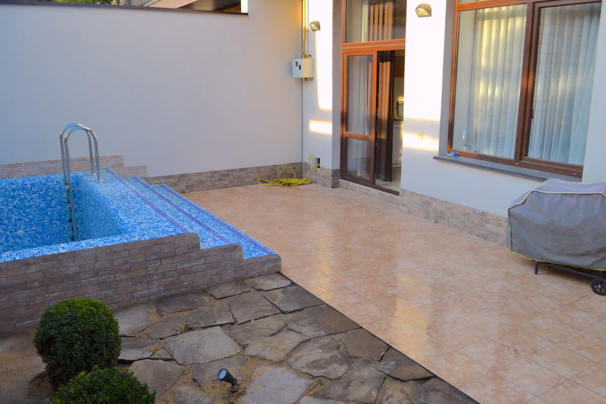 Una piscina con una escalera metálica, una parrilla cubierta y un camino de piedra que lleva a una puerta.