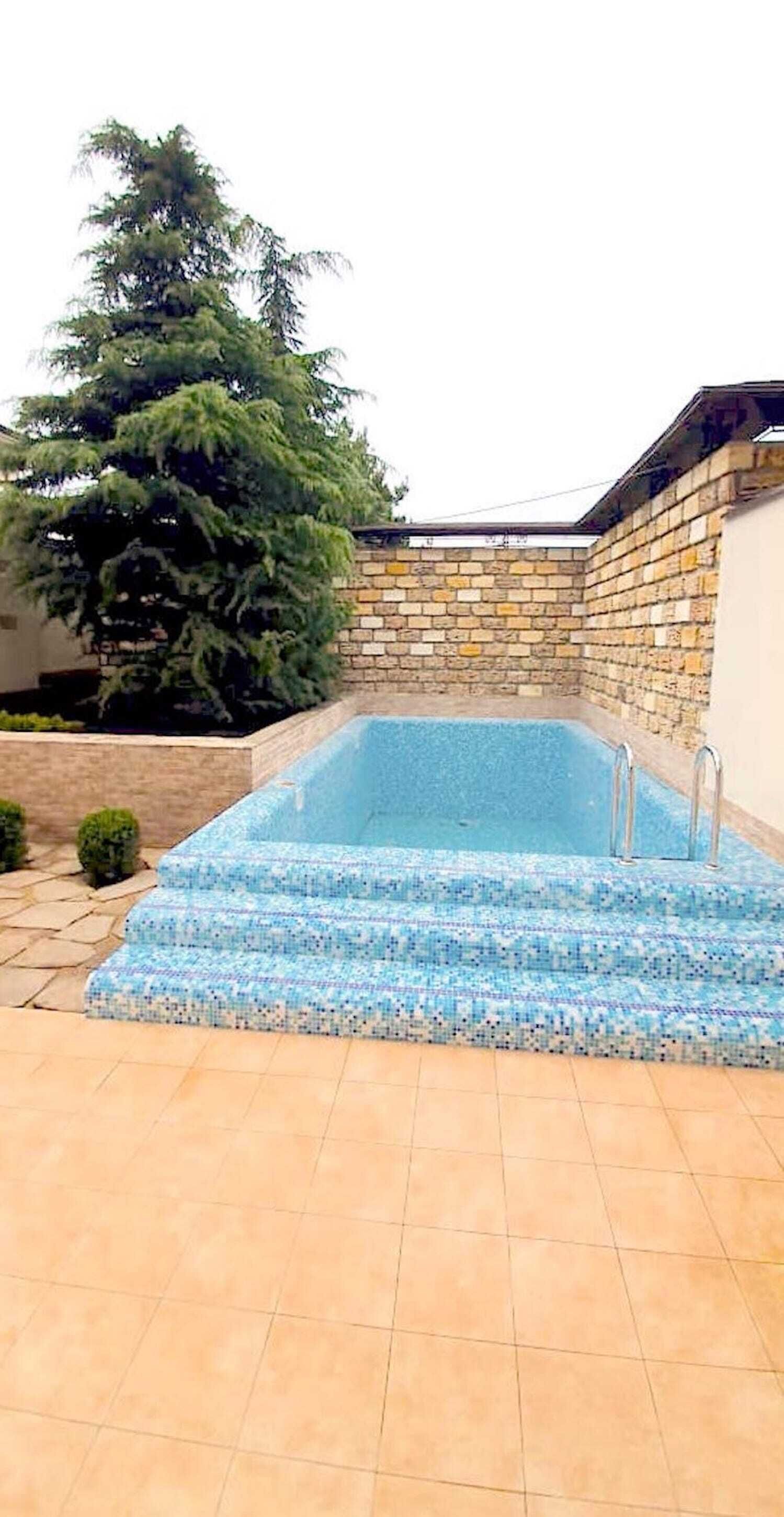 A rectangular swimming pool with a mosaic tile edge, surrounded by a stone wall and a tiled patio.