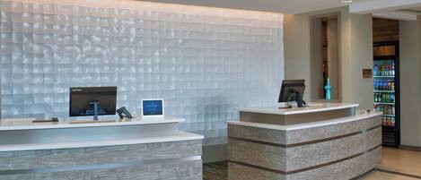 A reception area with two counters, one with a Lenovo monitor and a digital sign, and the other with a phone and a digital sign. There is a refrigerator stocked with beverages in the background.