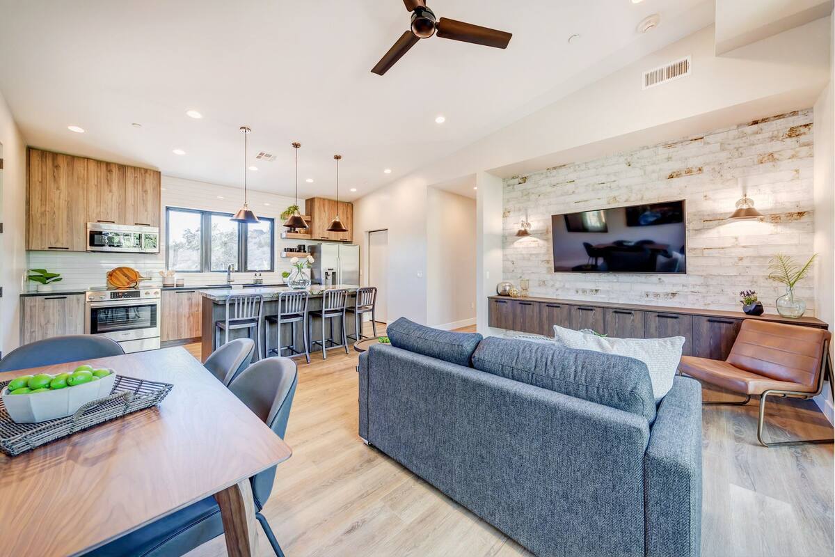 A modern kitchen with wooden cabinets, a dining area with a table and chairs, a gray sofa, and a TV mounted on a brick wall.