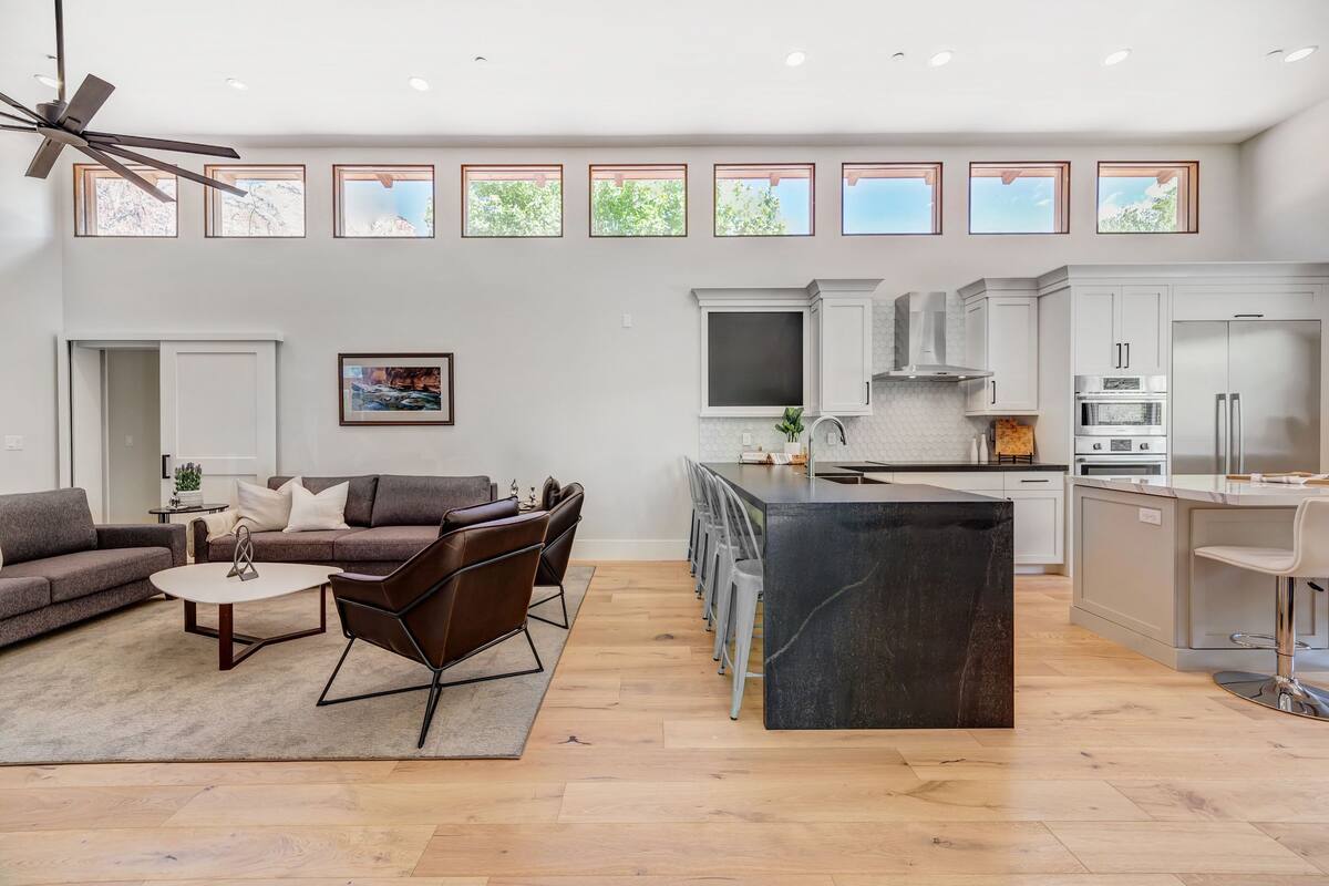 A modern living room with a sofa, coffee table, and chairs. There is a kitchen area with cabinets, a countertop, and a dining area with stools.