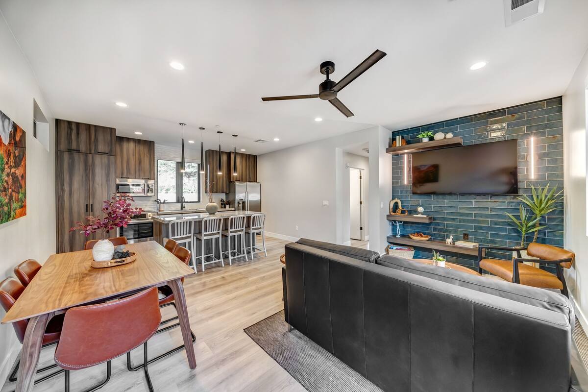 A modern kitchen with wooden cabinets, a dining area with a wooden table and chairs, a black leather sofa, and a ceiling fan.