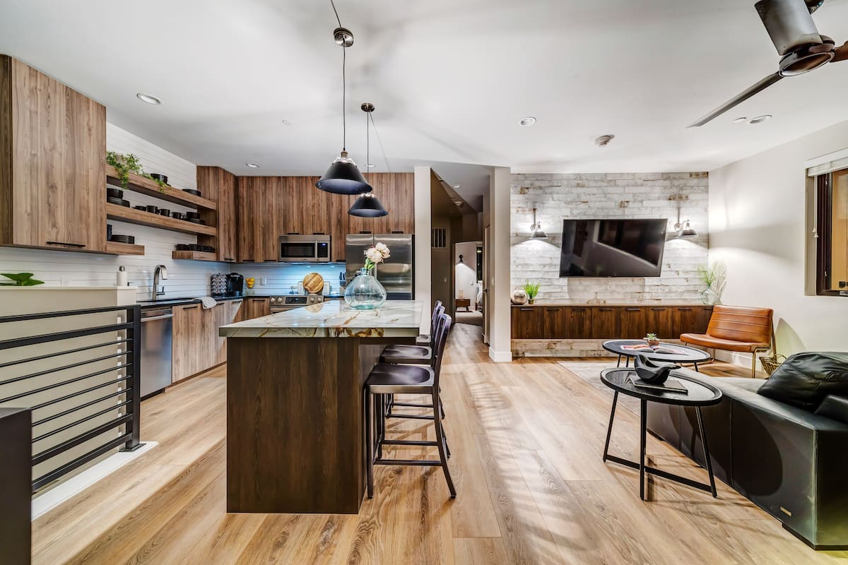 A modern kitchen with wooden cabinets, a central island, bar stools, and a dining area with a black sofa and round table.