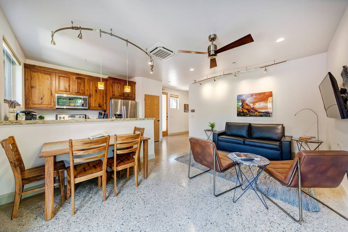 A modern kitchen with wooden cabinets, a stainless steel refrigerator, and a microwave. There is a dining area with a wooden table and chairs. A living room with a blue sofa and a coffee table is visible.