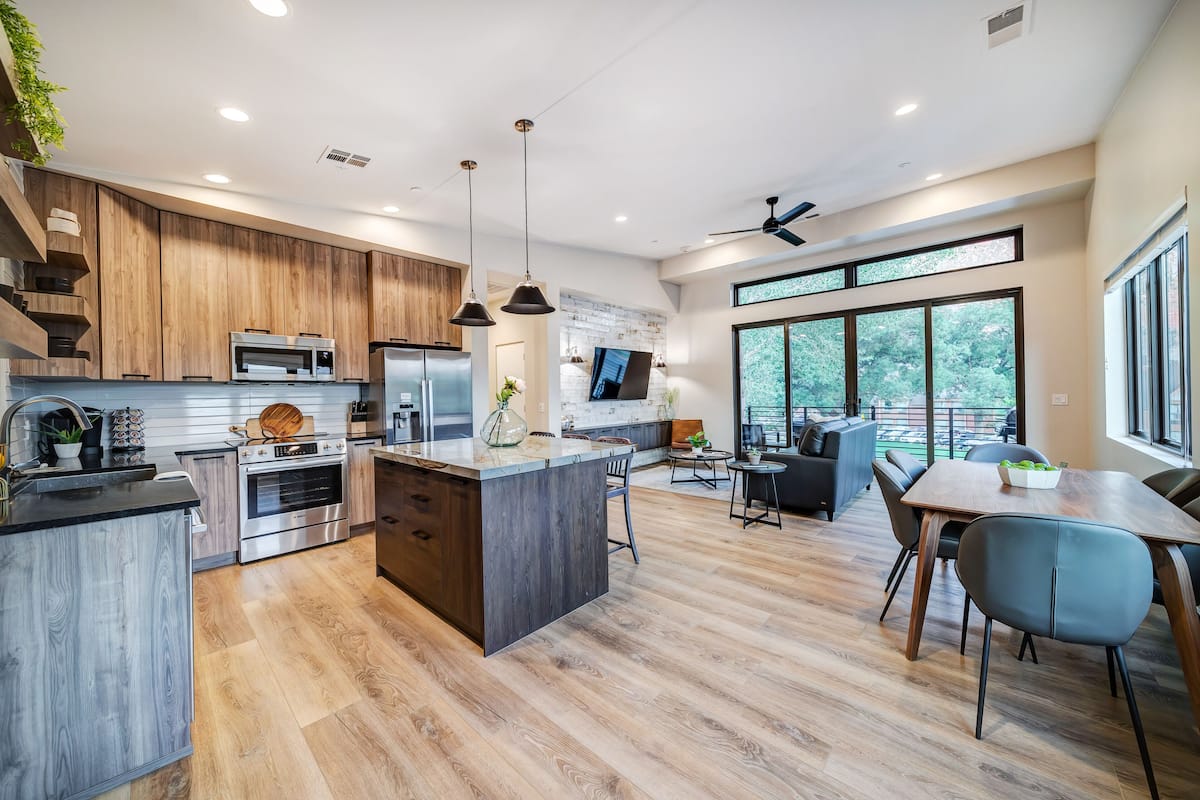A modern kitchen with wooden cabinetry, stainless steel appliances, and a central island. There is a dining area with a table and chairs, and large windows offering a view of the outdoors.