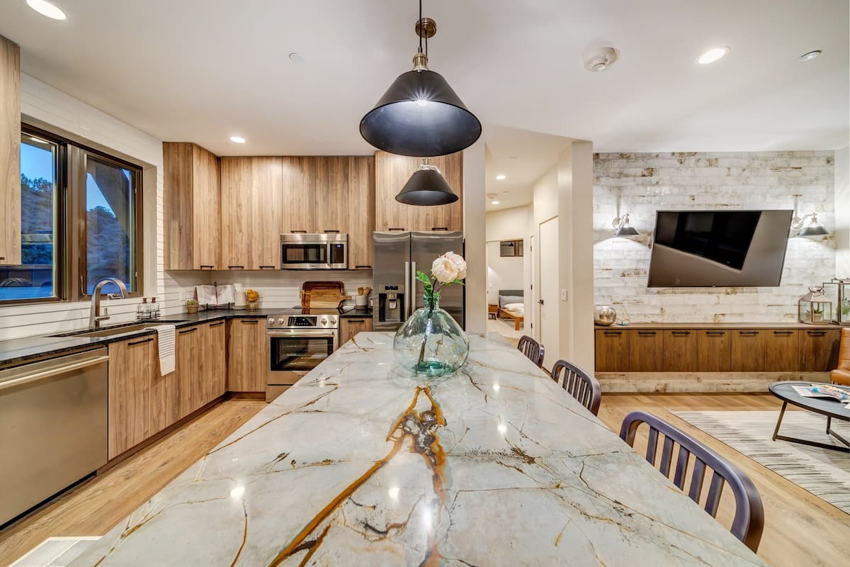 A modern kitchen with wooden cabinets, a marble island, and a large TV mounted on a brick wall.