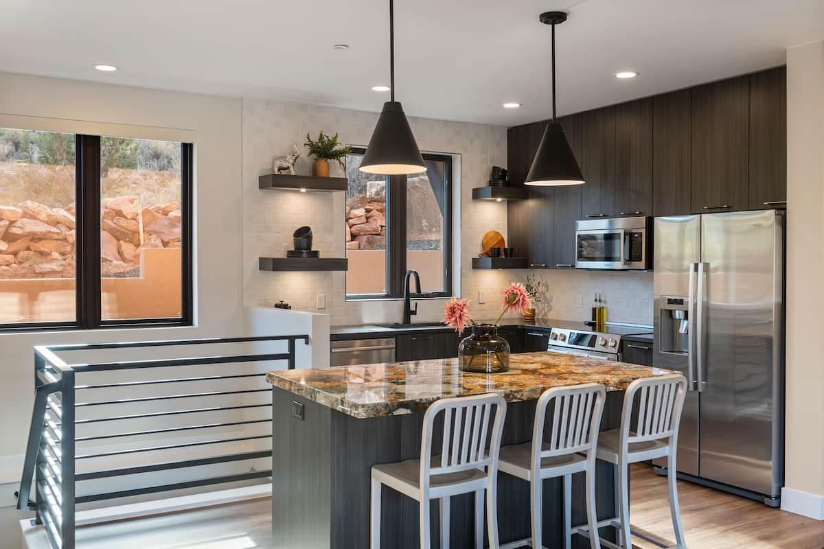 A modern kitchen with a large island, bar stools, dark wood cabinets, stainless steel appliances, and a large window with a view of rocks.