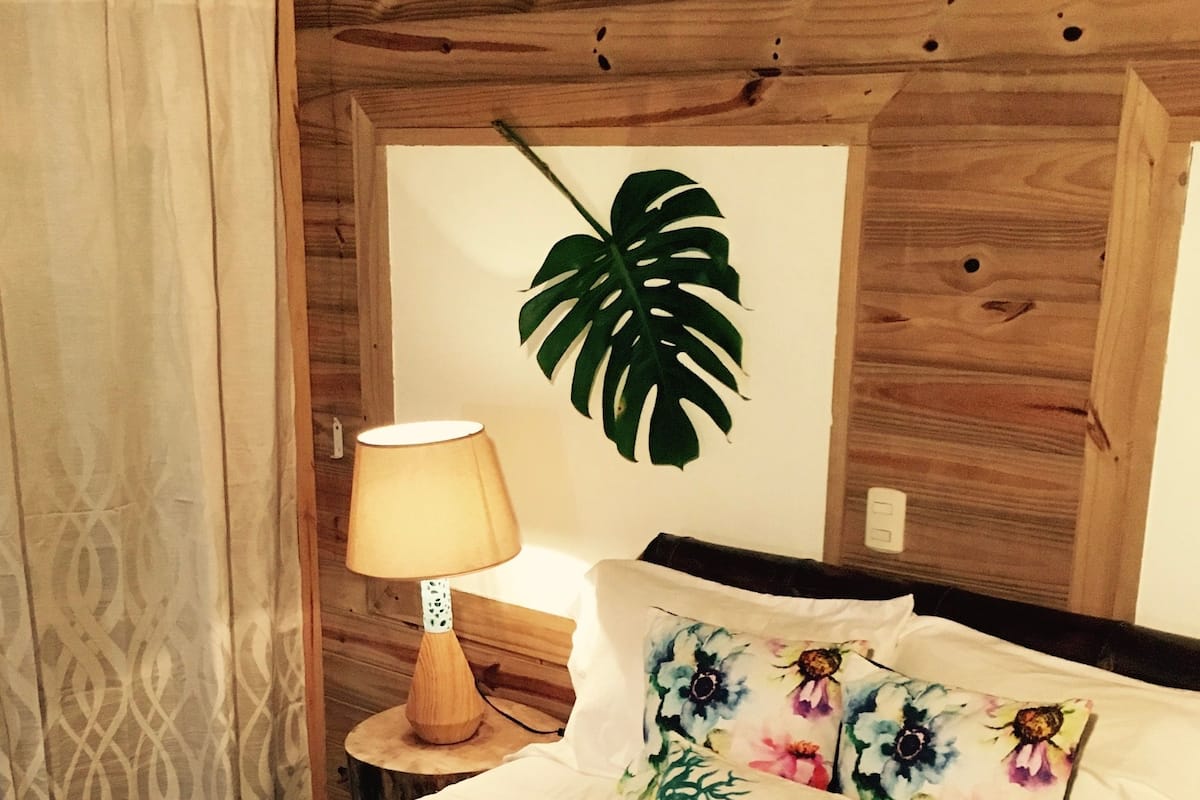 A bed with a white sheet, a green and white striped pillow, and a floral pillow. There is a wooden headboard, a nightstand with a lamp, and a wall with a large leaf decoration.