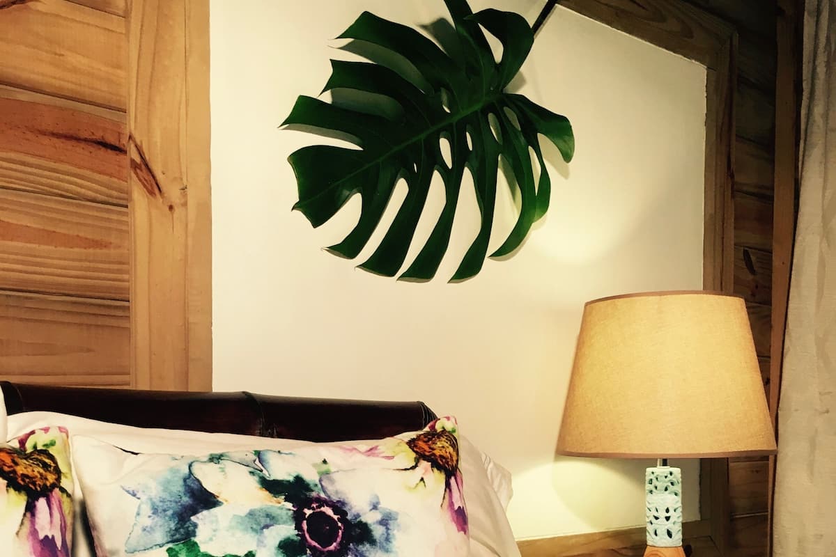 A bed with floral and tree-patterned pillows, a wooden bedside table with a lamp, and a large green leaf decoration on the wall.