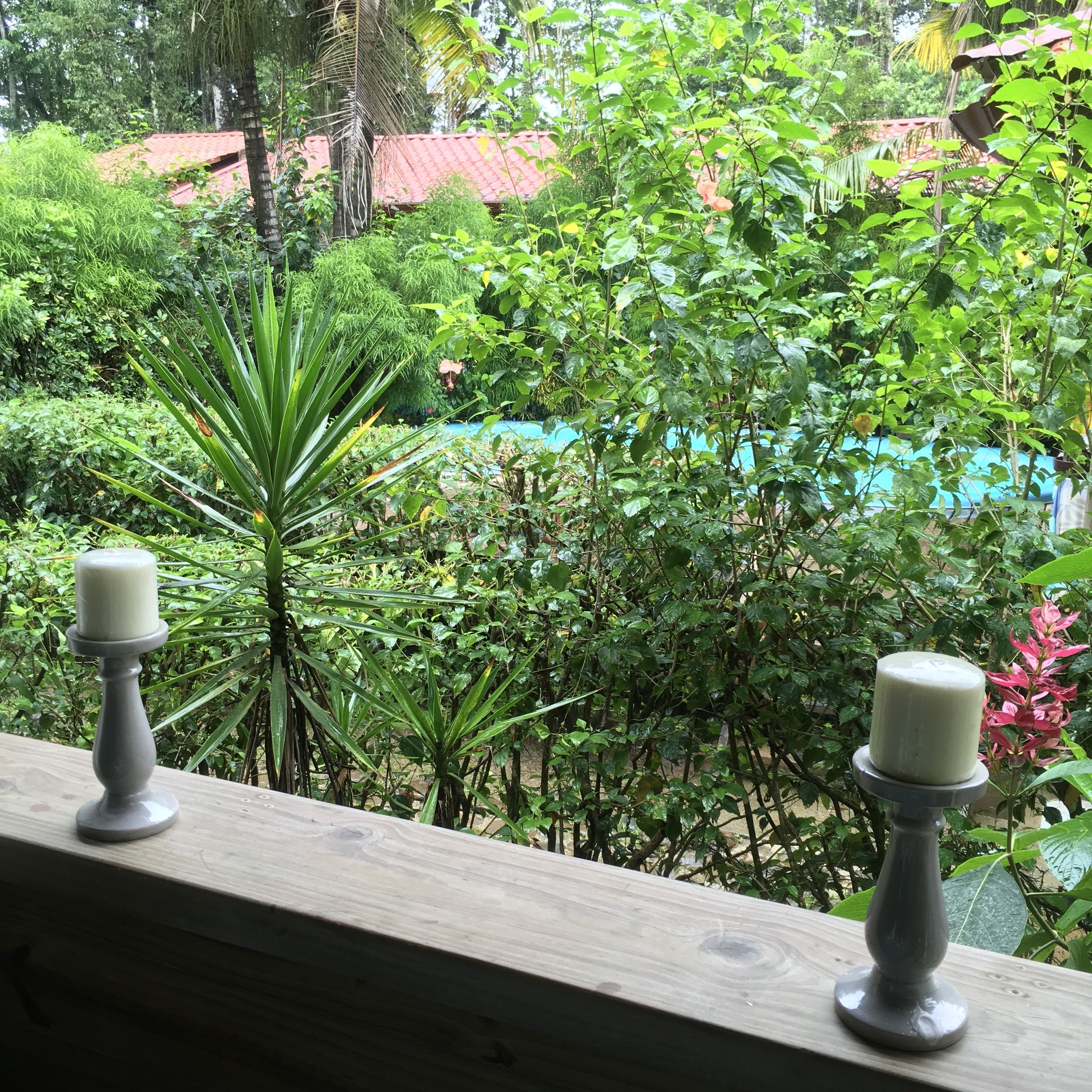 A wooden railing with two candle holders, surrounded by lush greenery and a red-roofed building in the background.