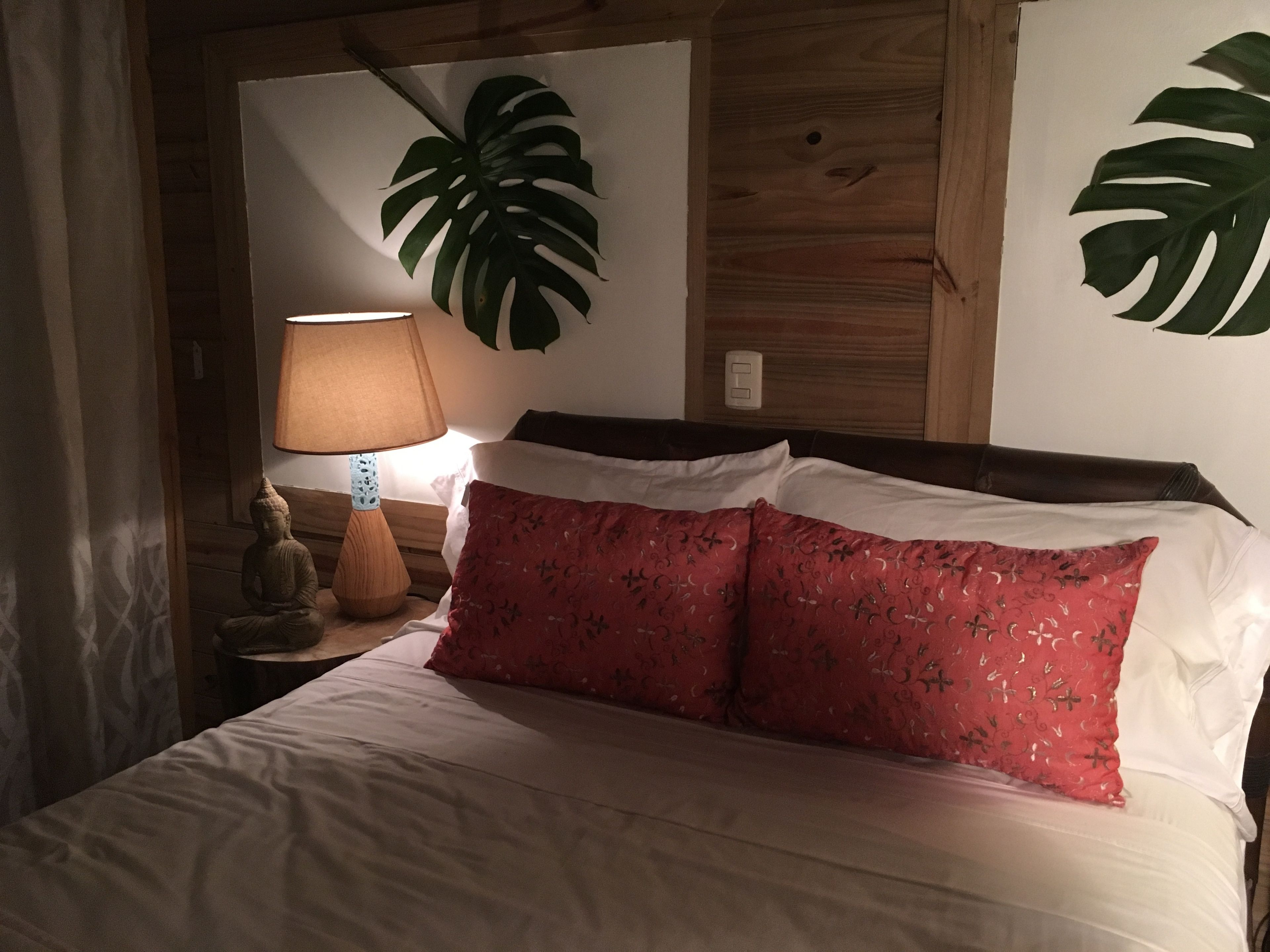 A bed with white linens and a red floral pillow, a wooden headboard, a wall-mounted lamp, and a Buddha statue on a nightstand.